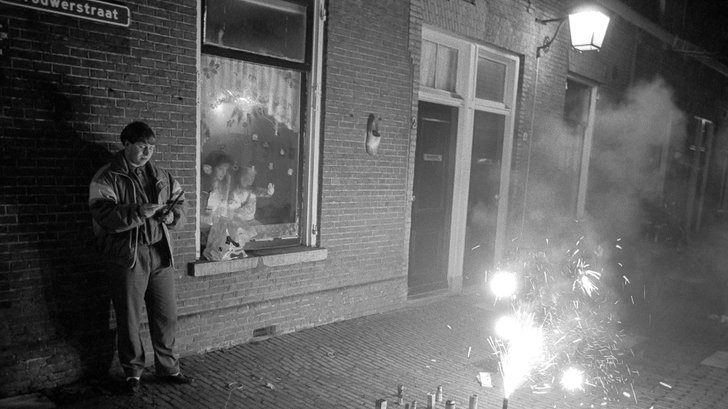 Een man steekt vuurwerk af tijdens Oud en Nieuw in de Brouwerstraat (Zeven Steegjes) in Utrecht, terwijl een vrouw en een kind door de ruit toekijken. 